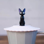 A close-up shot focuses on a small Jiji figurine sitting atop a pink scalloped silicone lid on a grey mug.