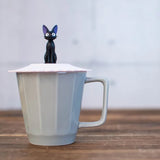 A Jiji figurine stands in the center of a scalloped Pink silicone mug lid on top of grey mug.
