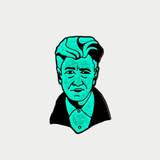 An enamel lapel pin depicting a green glowing bust illustration of filmmaker David Lynch with his signature swept-back white hair, a dark jacket, and a buttoned shirt against a plain gray background.