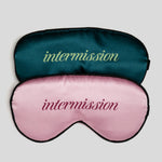 Two satin sleep masks stacked on a light gray background. The top mask is dark teal with the word 'intermission' in light green script, and the bottom mask is light pink with the word 'intermission' in dark pink script.