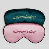 Two satin sleep masks stacked on a light gray background. The top mask is dark teal with the word 'intermission' in light green script, and the bottom mask is light pink with the word 'intermission' in dark pink script.