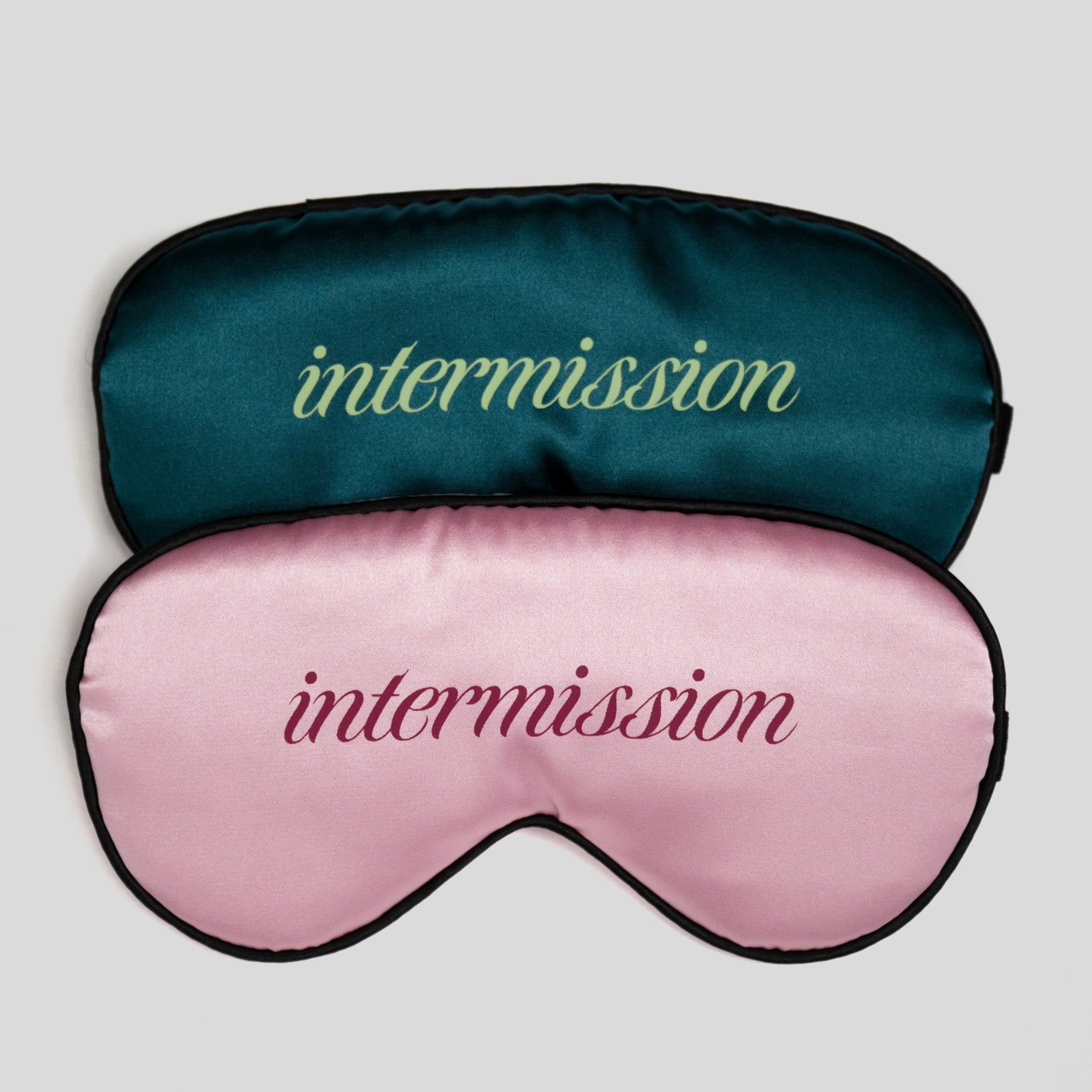 Two satin sleep masks stacked on a light gray background. The top mask is dark teal with the word 'intermission' in light green script, and the bottom mask is light pink with the word 'intermission' in dark pink script.