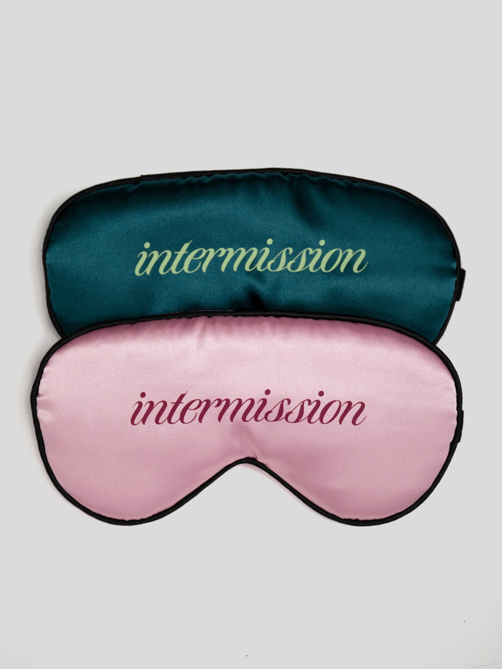 Two satin sleep masks stacked on a light gray background. The top mask is dark teal with the word 'intermission' in light green script, and the bottom mask is light pink with the word 'intermission' in dark pink script.