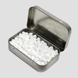 An open silver metal tin filled with small white round tablets, photographed on a light gray background.