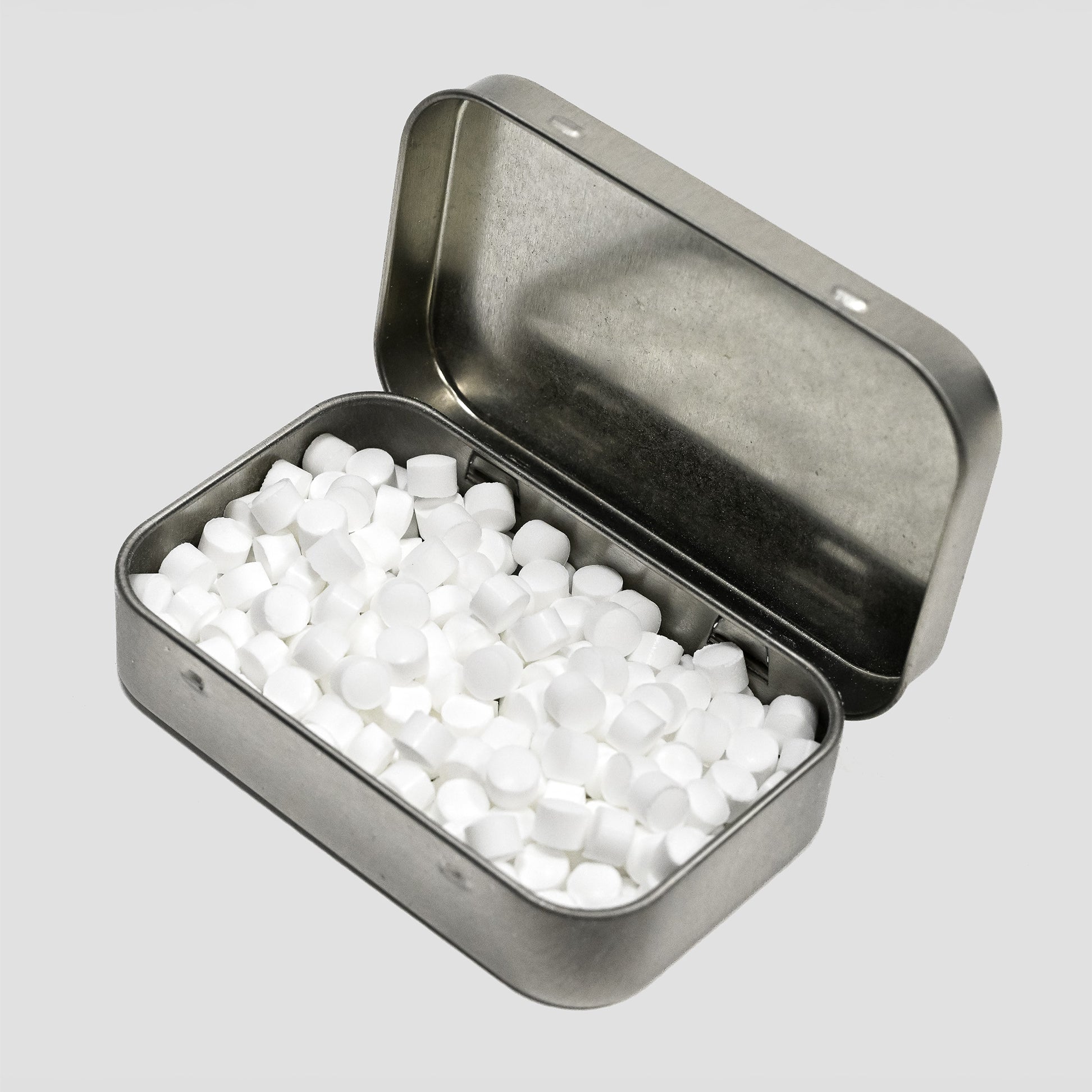 An open silver metal tin filled with small white round tablets, photographed on a light gray background.