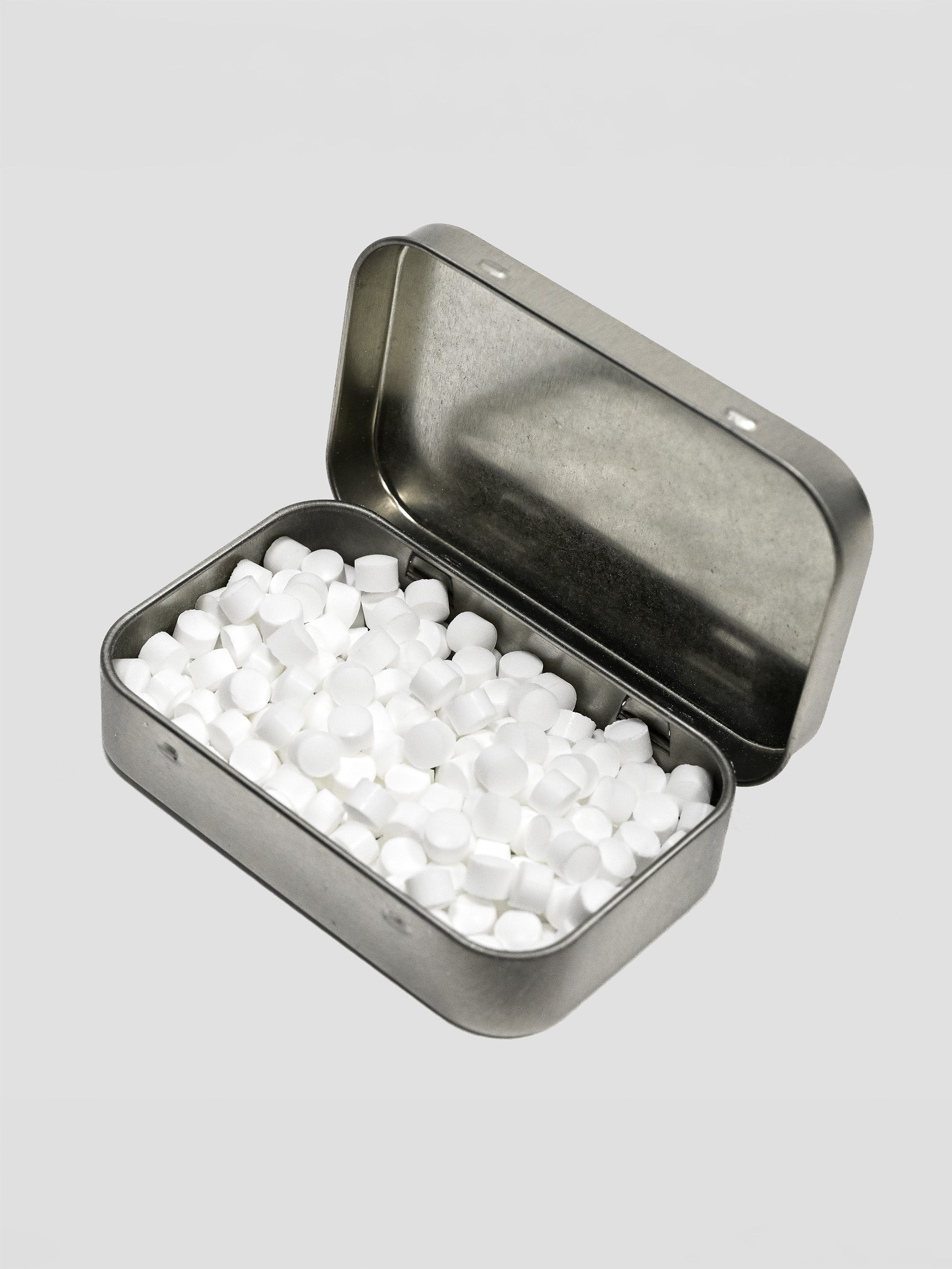 An open silver metal tin filled with small white round tablets, photographed on a light gray background.
