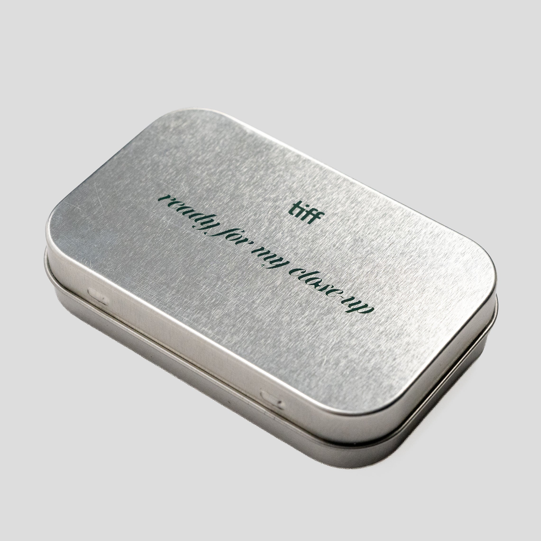 A rectangular silver metal tin with rounded corners, featuring the words 'ready for my close-up' and 'tiff' printed on the lid, set against a solid light gray background.