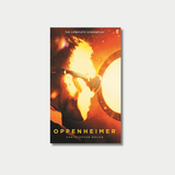 The book cover for Christopher Nolan's "Oppenheimer: The Complete Screenplay" shows the silhouette of a man wearing protective goggles as he is intensely illuminated by a fiery, golden nuclear blast, centered against a light gray background.