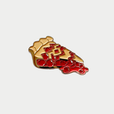 An enamel lapel pin shaped like a slice of cherry pie with glossy red cherry filling, a golden lattice crust, and gold trim against a plain light gray background.