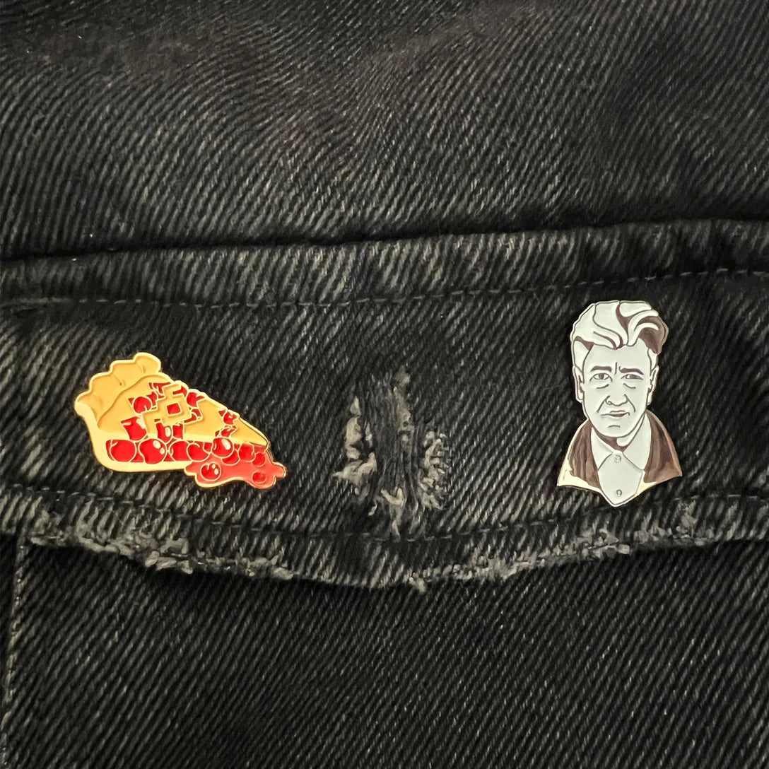 Two enamel lapel pins—one of a cherry pie slice on the left and one of David Lynch on the right—pinned to the pocket flap of a black denim jacket.