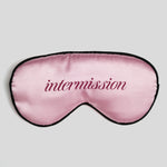 A pink satin sleep mask with black trim and the word 'intermission' embroidered in dark pink script across the center, shown on a light gray background.