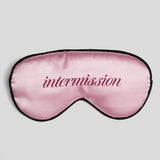 A pink satin sleep mask with black trim and the word 'intermission' embroidered in dark pink script across the center, shown on a light gray background.