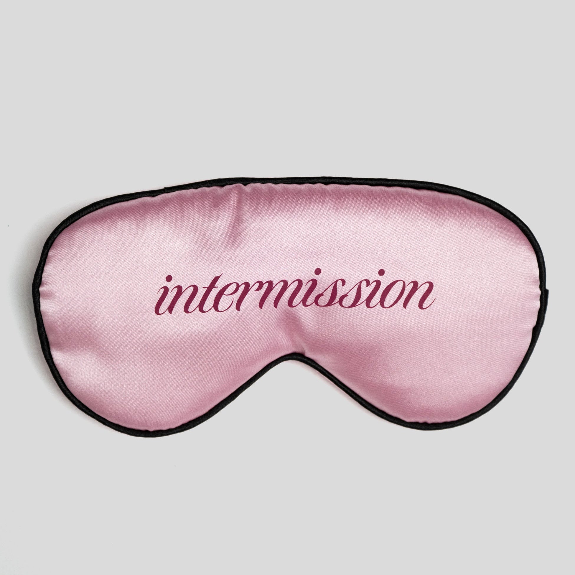 A pink satin sleep mask with black trim and the word 'intermission' embroidered in dark pink script across the center, shown on a light gray background.