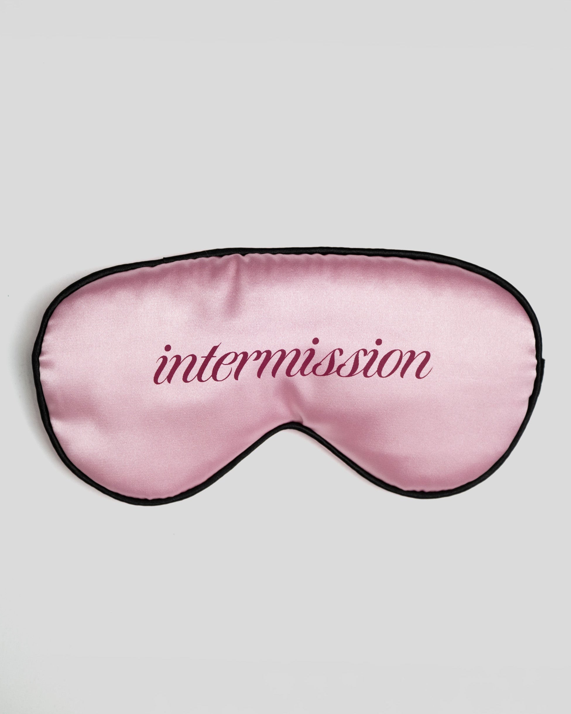 A pink satin sleep mask with black trim and the word 'intermission' embroidered in dark pink script across the center, shown on a light gray background.