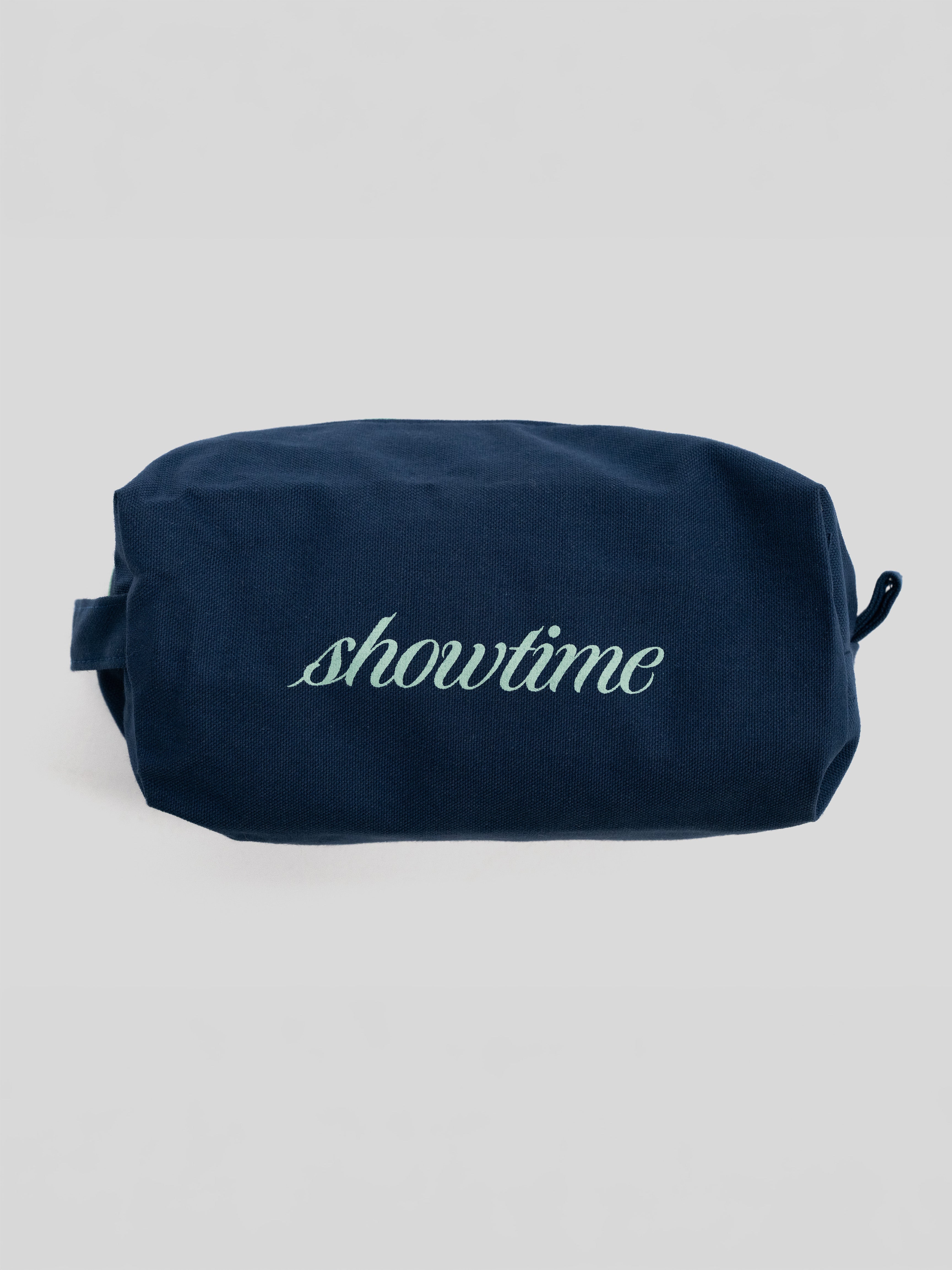 A navy blue canvas cosmetic pouch with the word 'showtime' embroidered in light green script across the front, set against a plain light gray background.