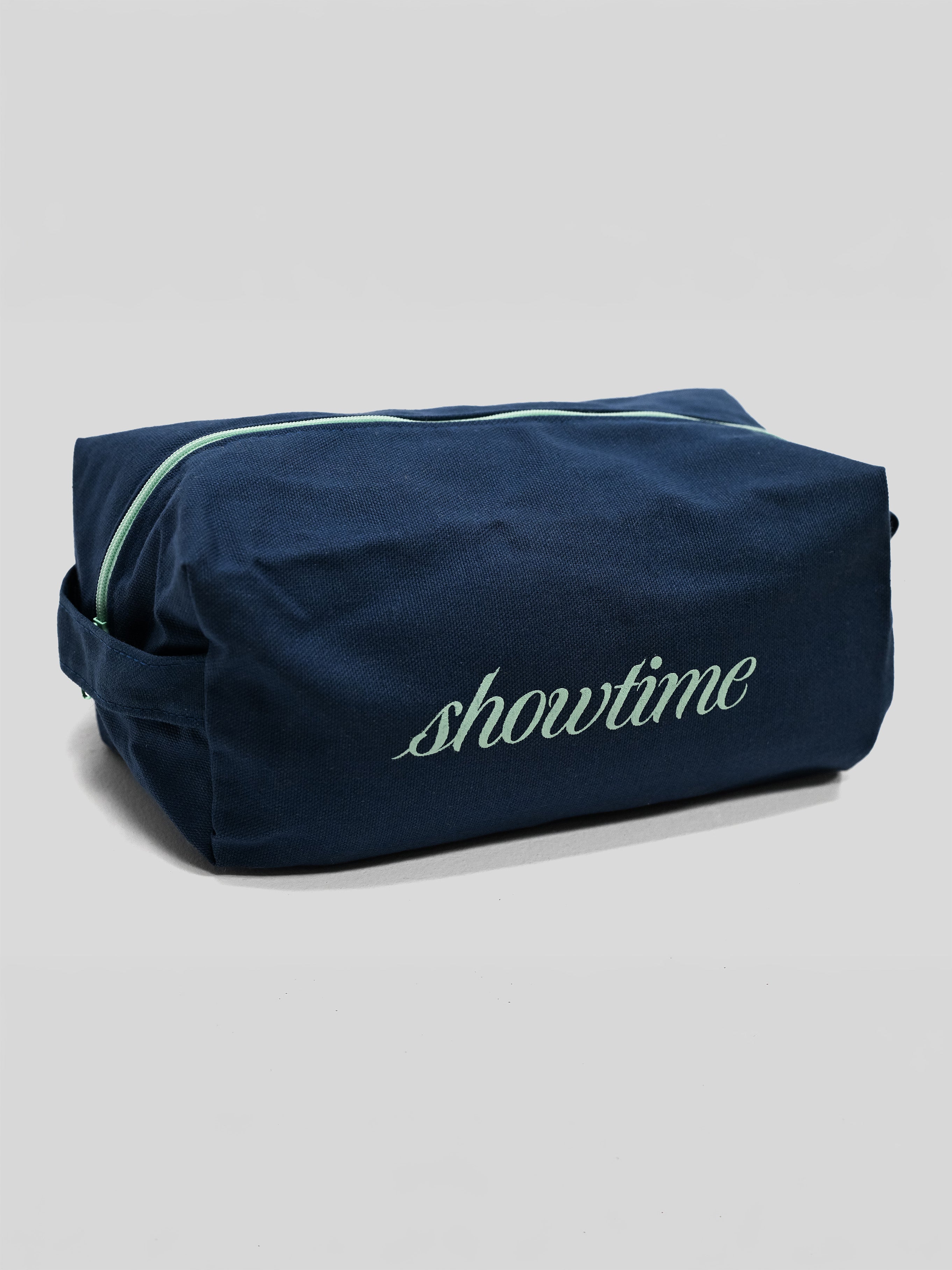A navy blue toiletry bag with a mint green zipper and 'showtime' embroidered in mint green on the front, set against a light gray background.