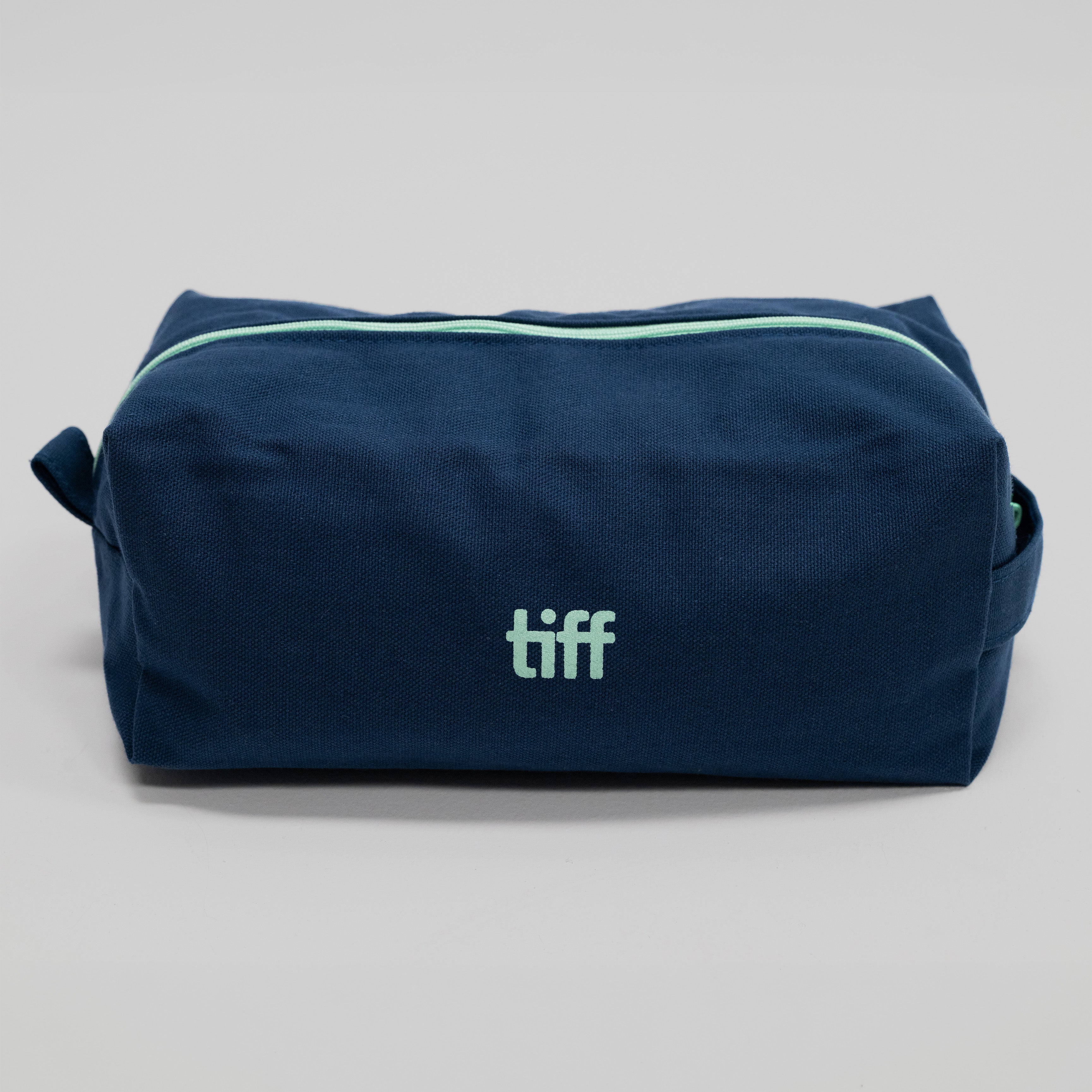 A navy blue canvas toiletry bag with a mint green zipper and a mint green 'tiff' logo on the front, photographed on a neutral light gray background.