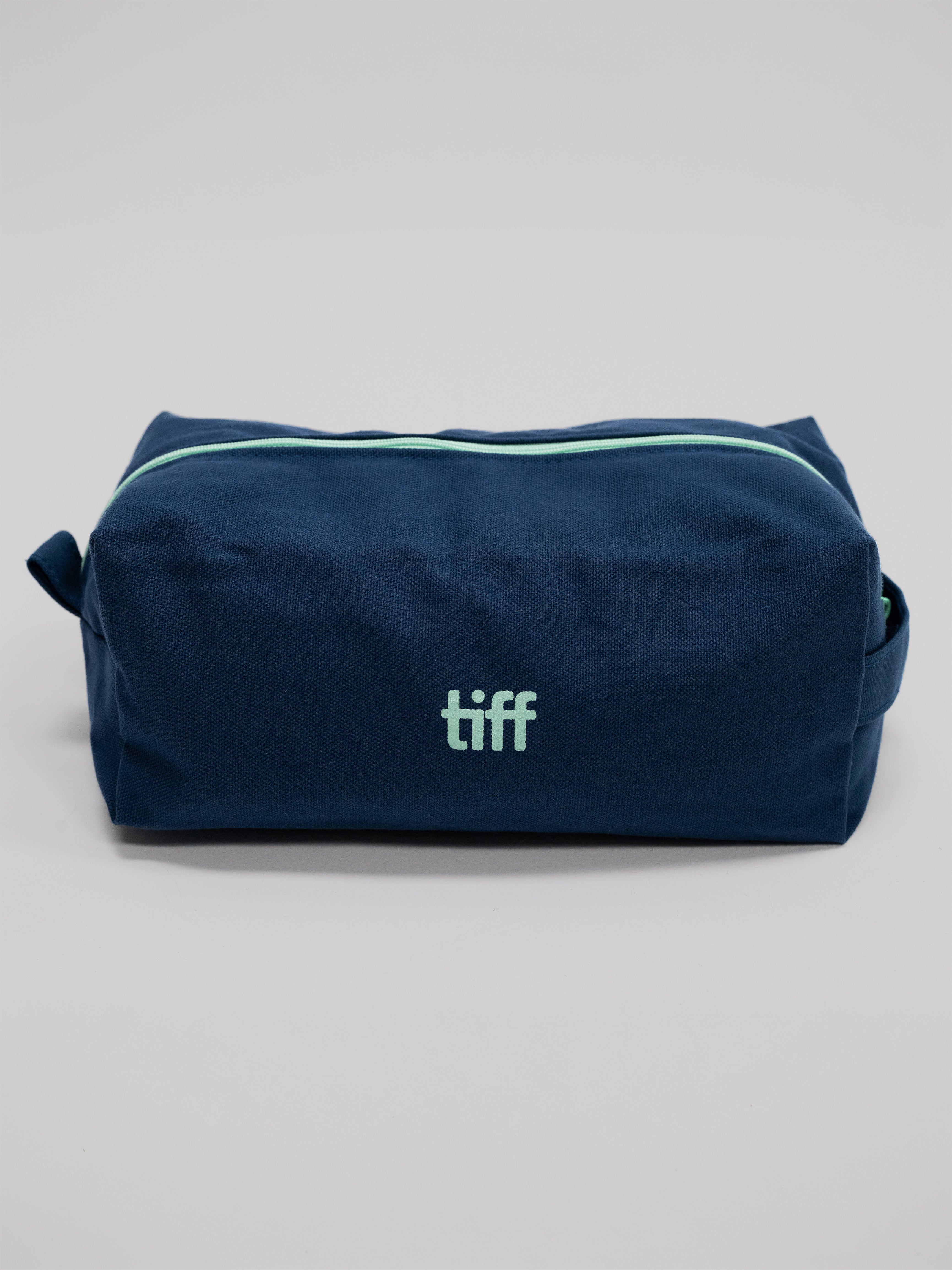 A navy blue canvas toiletry bag with a mint green zipper and a mint green 'tiff' logo on the front, photographed on a neutral light gray background.