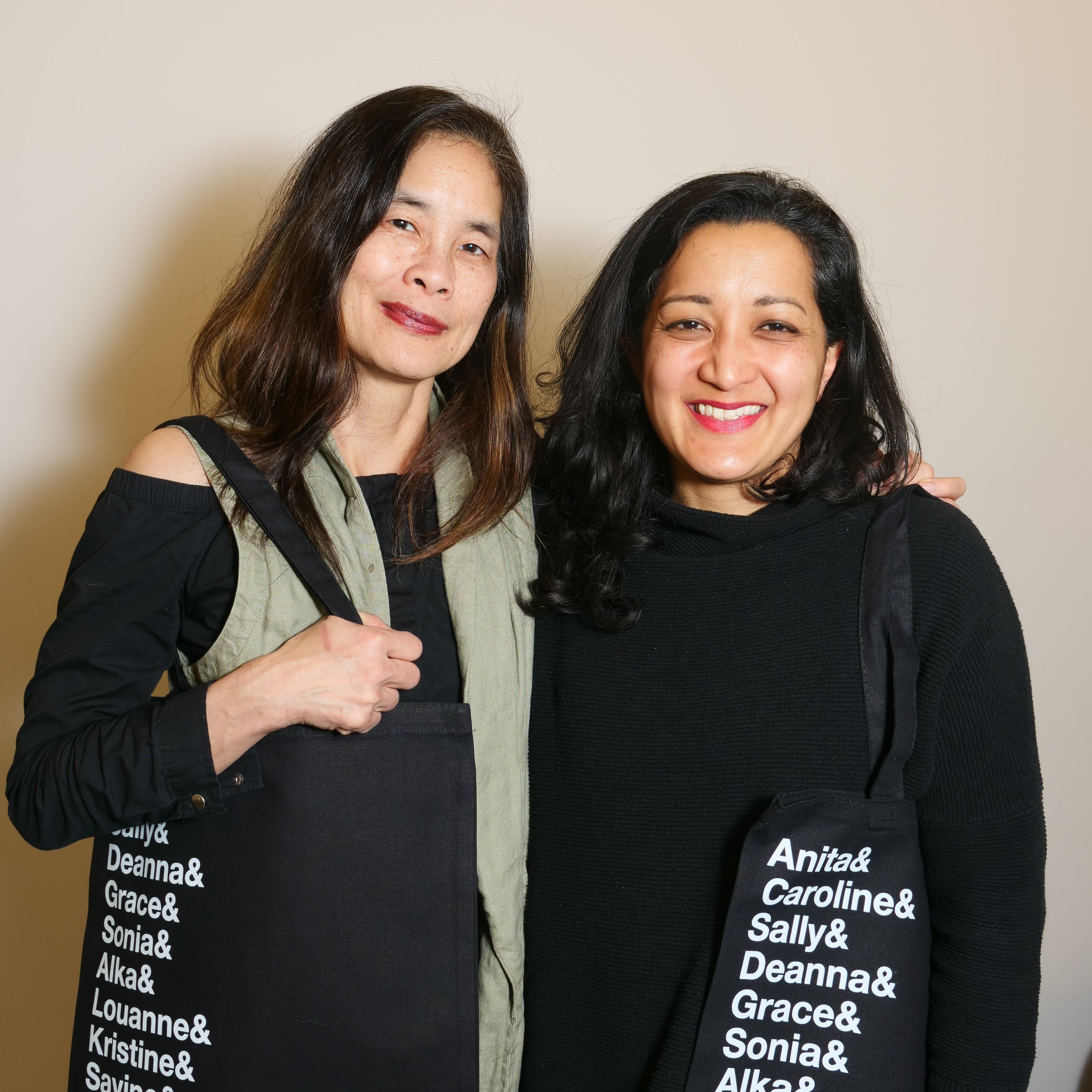 Two people in black clothing smiling at the camera carrying black tote bags printed with a list of names in white text