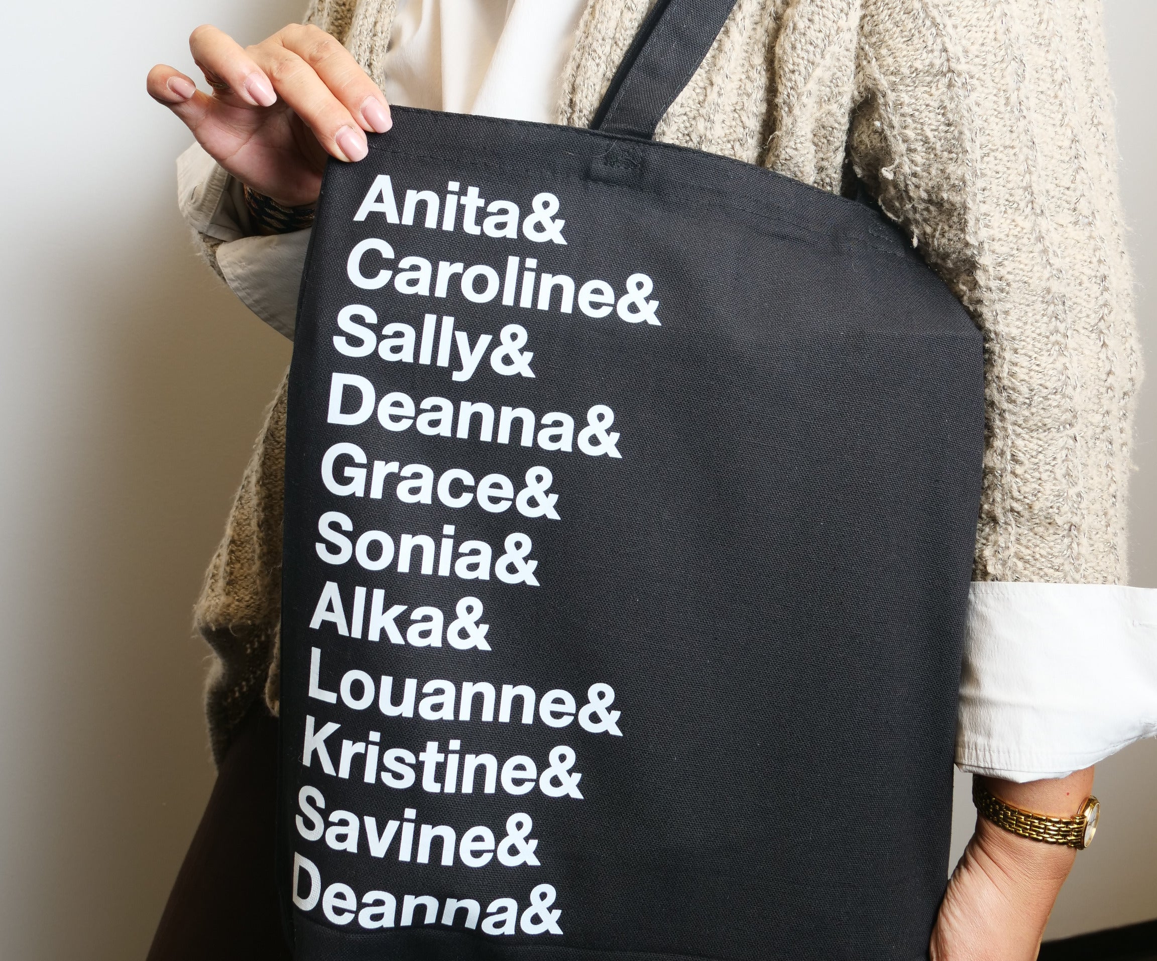 Person in a cream sweater holding a black tote bag with white text listing multiple names, standing against a plain wall