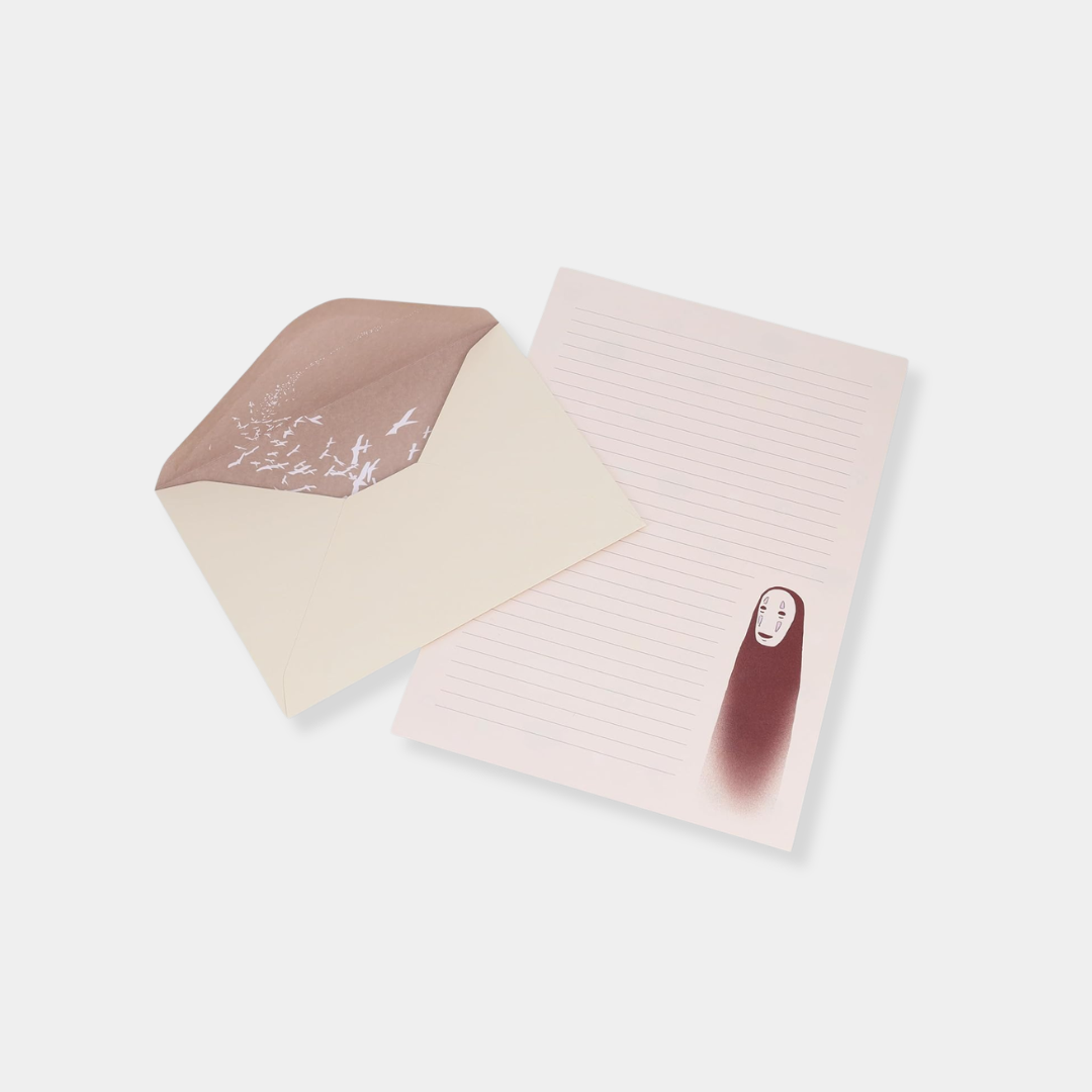 An open cream-colored envelope with a bird-patterned interior liner sits next to a sheet of light pink lined stationery featuring an illustration of the character No-Face from "Spirited Away."