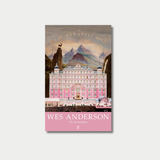 Book cover featuring a symmetrical, pastel pink illustration of the Grand Budapest Hotel set against a backdrop of rugged mountains, with a prominent pink banner at the bottom reading "WES ANDERSON The Screenplays.