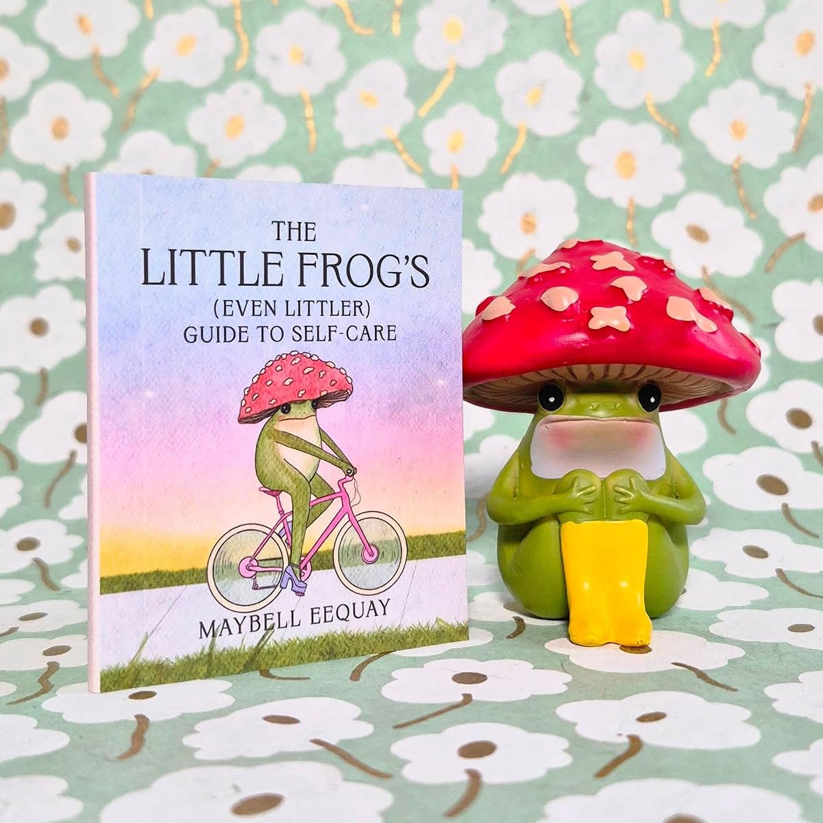 A small book titled "The Little Frog's (Even Littler) Guide to Self-Care" stands next to a matching green frog figurine wearing a red mushroom cap, both set against a light green floral background.
