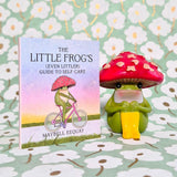 A small book titled "The Little Frog's (Even Littler) Guide to Self-Care" stands next to a matching green frog figurine wearing a red mushroom cap, both set against a light green floral background.