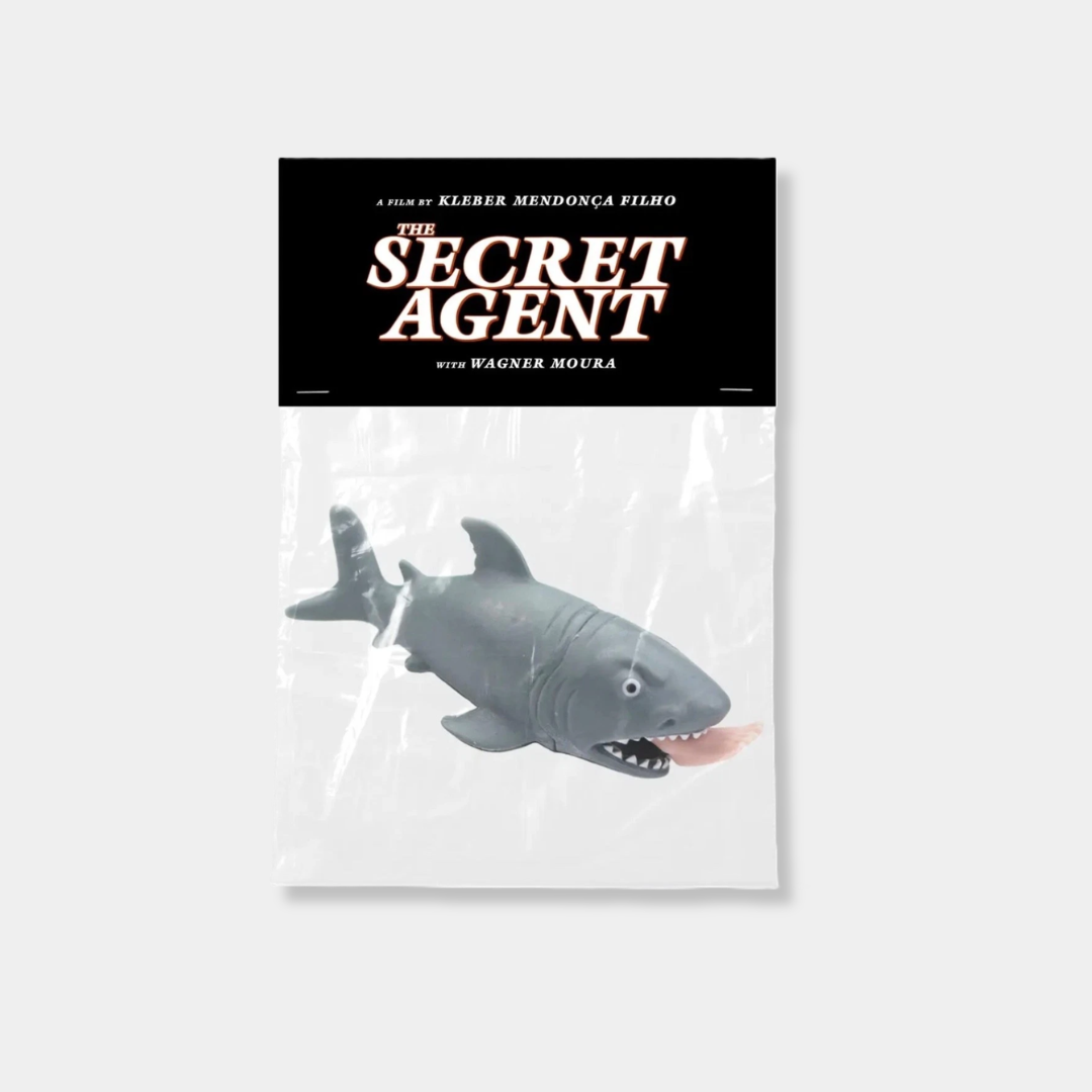 A grey rubber shark toy with a human leg protruding from its mouth is packaged in a clear plastic bag with a black header card reading "The Secret Agent," a film by Kleber Mendonça Filho.