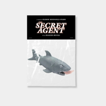 A grey rubber shark toy with a human leg protruding from its mouth is packaged in a clear plastic bag with a black header card reading "The Secret Agent," a film by Kleber Mendonça Filho.