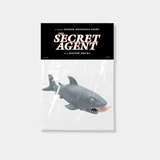 A grey rubber shark toy with a human leg protruding from its mouth is packaged in a clear plastic bag with a black header card reading "The Secret Agent," a film by Kleber Mendonça Filho.