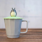 A grey fluted ceramic mug sits on a wooden surface, topped with a green silicone lid and a small Totoro figure.