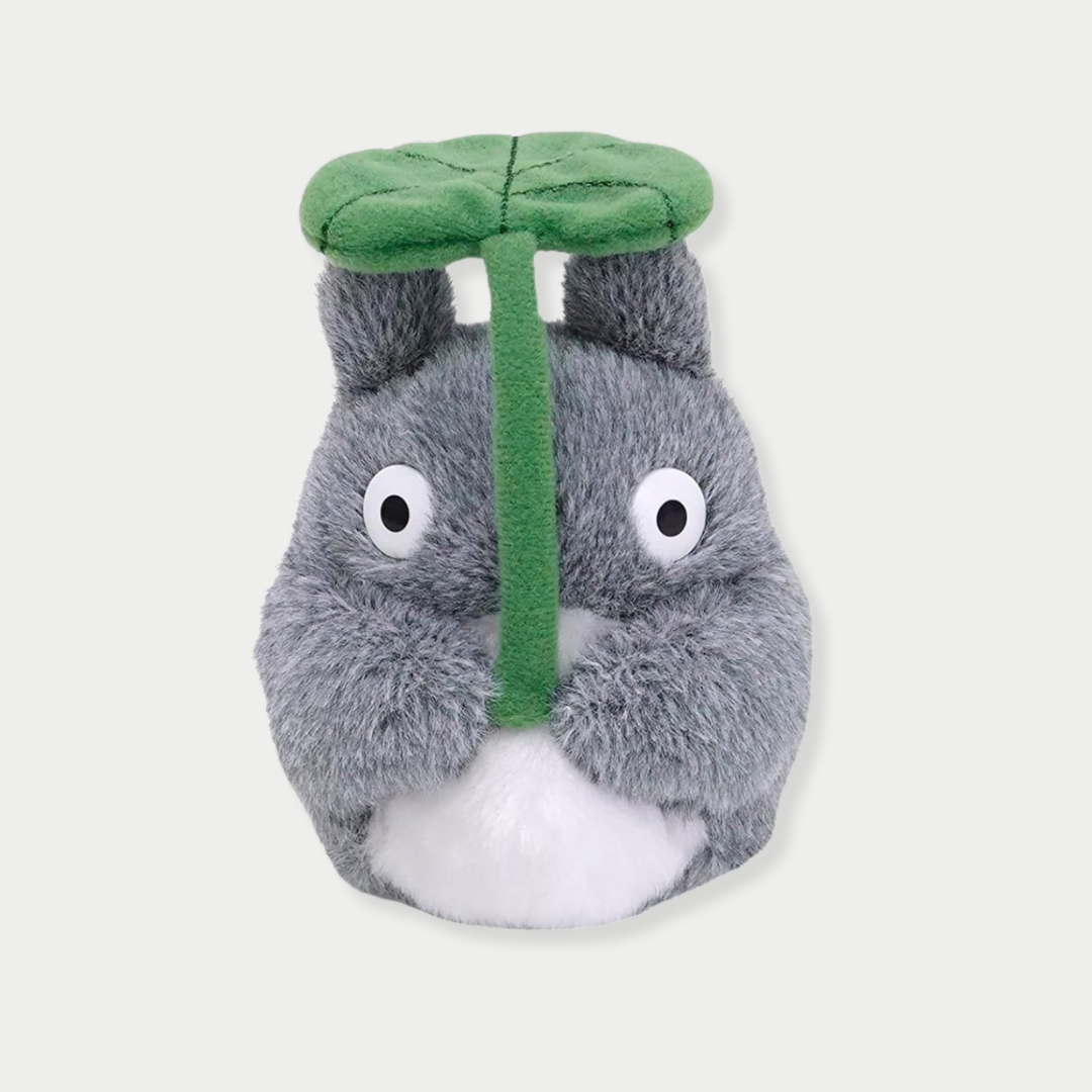 A fluffy gray Totoro plush toy holds a large green leaf overhead like an umbrella.