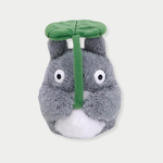 A fluffy gray Totoro plush toy holds a large green leaf overhead like an umbrella.