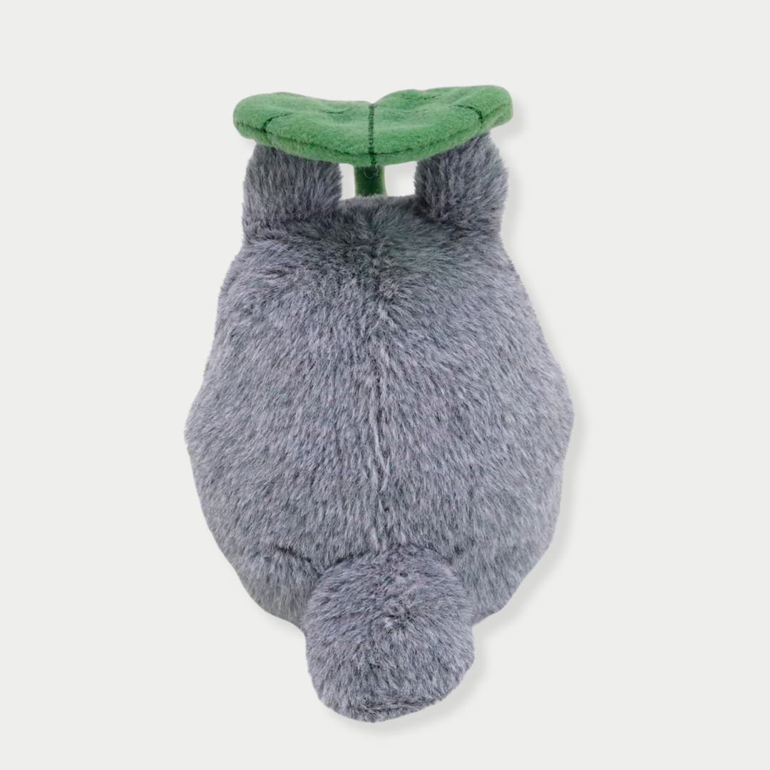 Back view of fluffy gray Totoro plush toy holding a large green leaf overhead like an umbrella.


