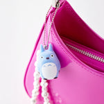 A light blue, flocked Totoro keychain attached to the silver zipper of a vibrant pink handbag.