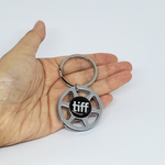 Photo of a hand holding a silver toned keychain that looks like a miniature film reel with the tiff logo in the centre of it.